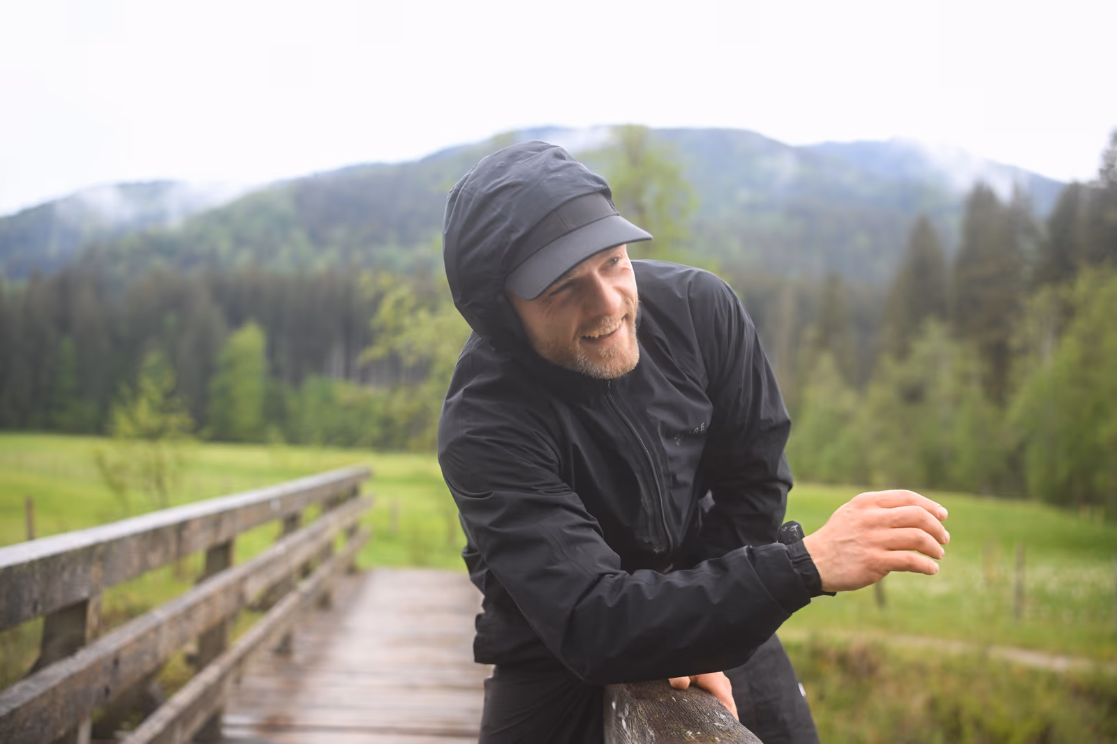 Aaron Schwägerl an ein Brückengeländer gelehnt, lächelnd, Alpenpanorama im Hintergrund