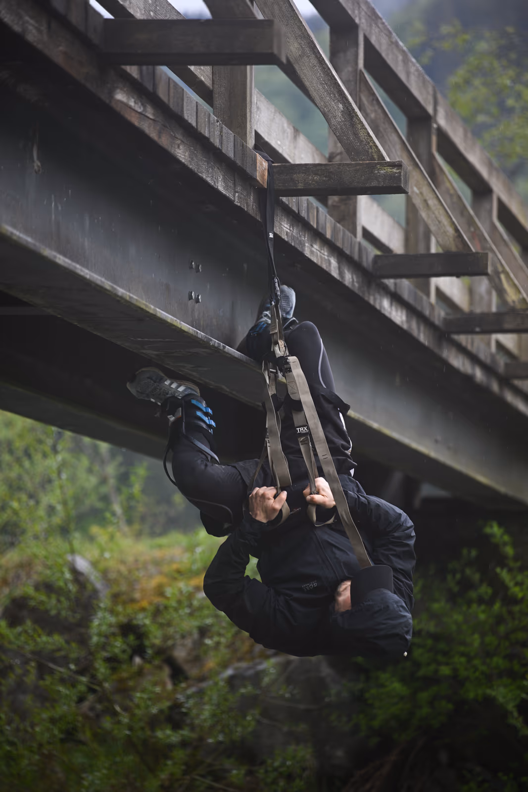 Aaron Schwägerl schwingt im TRX-Band unter einer Holzbrücke, Füße an der Brücke, Körper frei hängend